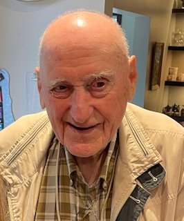 Front-facing portrait of an elderly man indoors, wearing a beige jacket and plaid shirt, smiling at the camera.