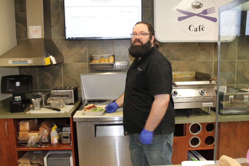 Chef with a beard and glasses wears a black shirt and blue gloves, preparing a salad at a cafe kitchen counter.