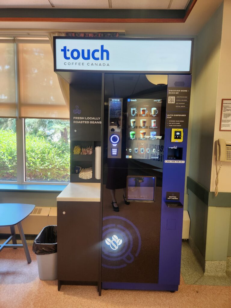 Vending coffee kiosk labeled 'touch COFFEE CANADA' with drink selections on a touchscreen panel in a cafe corner.